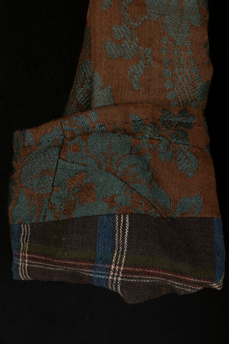 Handsydd bröllopsklänning i jacquardväv med grönt växtmönster mot brun bakgrund. Främre knäppning med nio hyskor, högknäppt och enkel halsring. Hellång ärm och kjol. Foder i ofärgat- respektive rutmönstrat linnetyg. Visst bruksslitage.