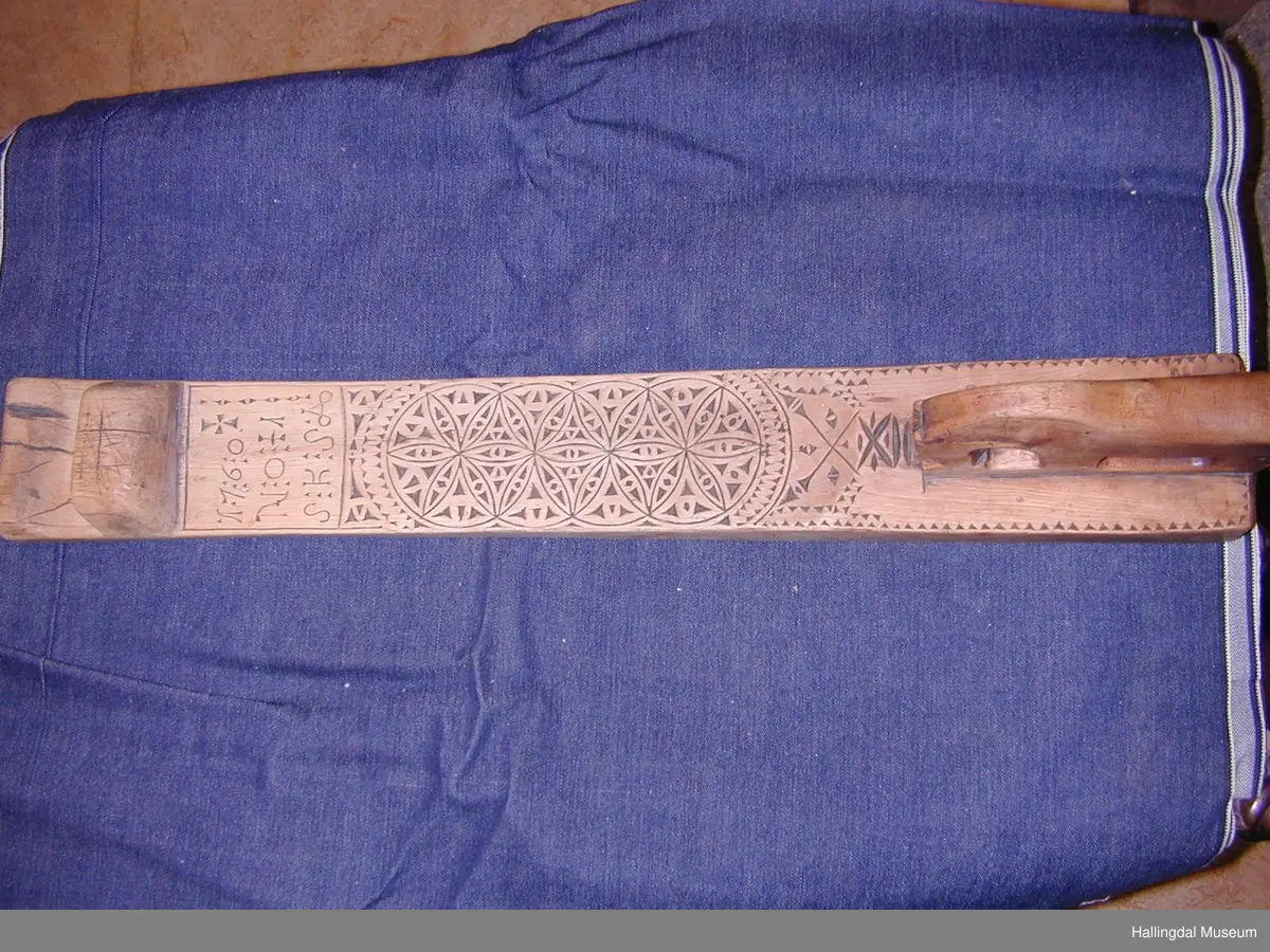 Mangletre, skore. Rektangulært tverrsnitt i sjølve fjøla, litt boga på undersida. På oversida skoren ornamentikk. På framenden på fjøla skore ein slags krok til å halde i. I attaste enden skore ein hest, felt ned i fjøla. Ytst i denne enden ei lærrreim festa med to naglar.