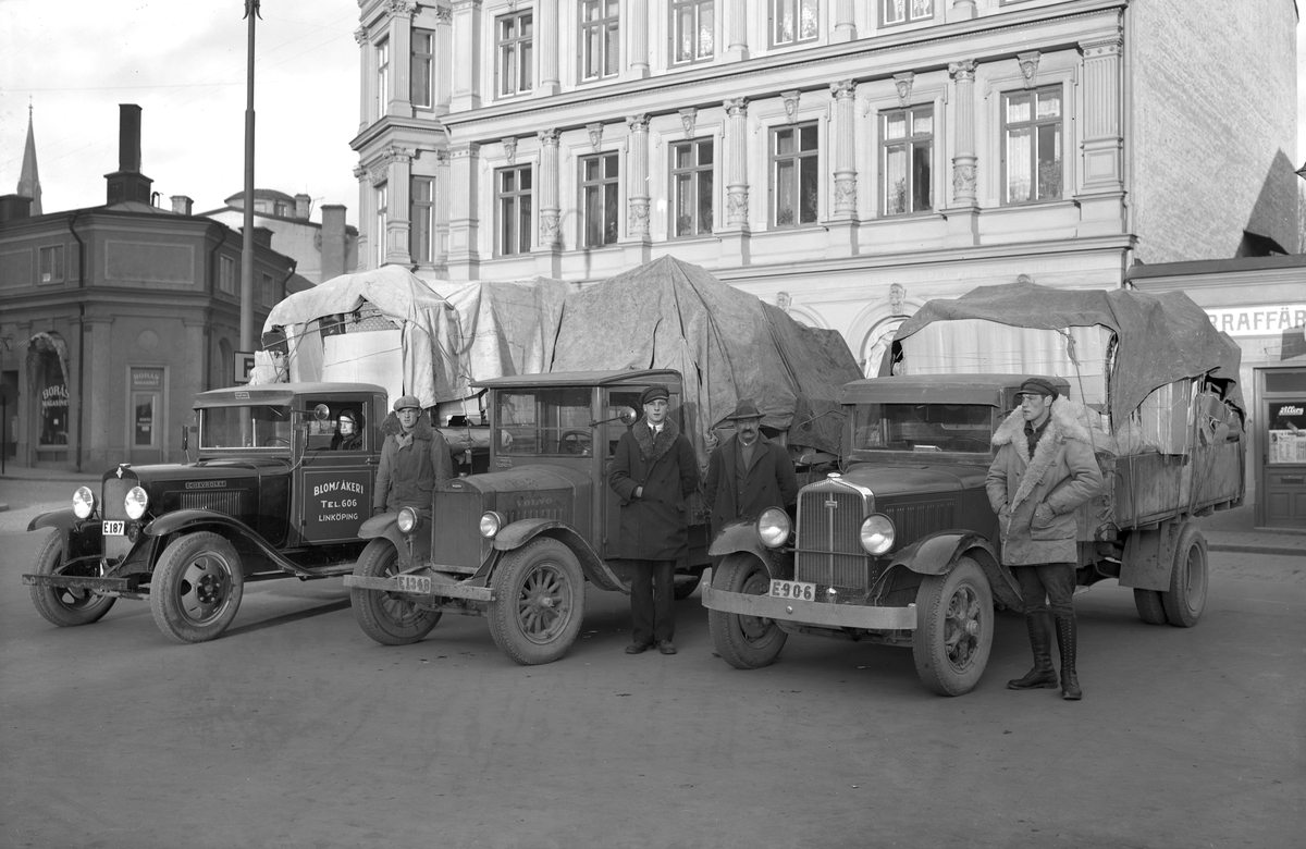 Herman Blom grundade Bloms Åkeri år 1919 som ett häståkeri, men redan 1924 köpte företaget in sin första lastbil och blev pionjärer inom fjärrtrafik. Nu är året 1932 och Herman med söner har ställt upp firmans lastbilsflotta på Trädgårdstorget i Linköping för reklamfotografering. Från vänster ses Ivan Blom vid en Chevrolet från 1930, Gösta och Herman Blom vid en Volvo av 1928 års modell samt Evert Blom vid en Studebaker från 1930. Kvinnan som skymtar i hytten har identifierats som Ivans fru Anna.