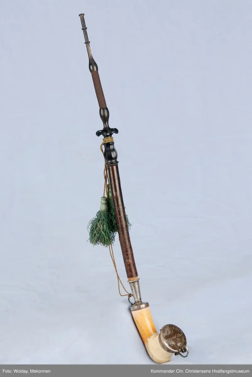 Langpipe med mellomstykke (holk) og lokk i sølv. Grønn snor med to dusker.