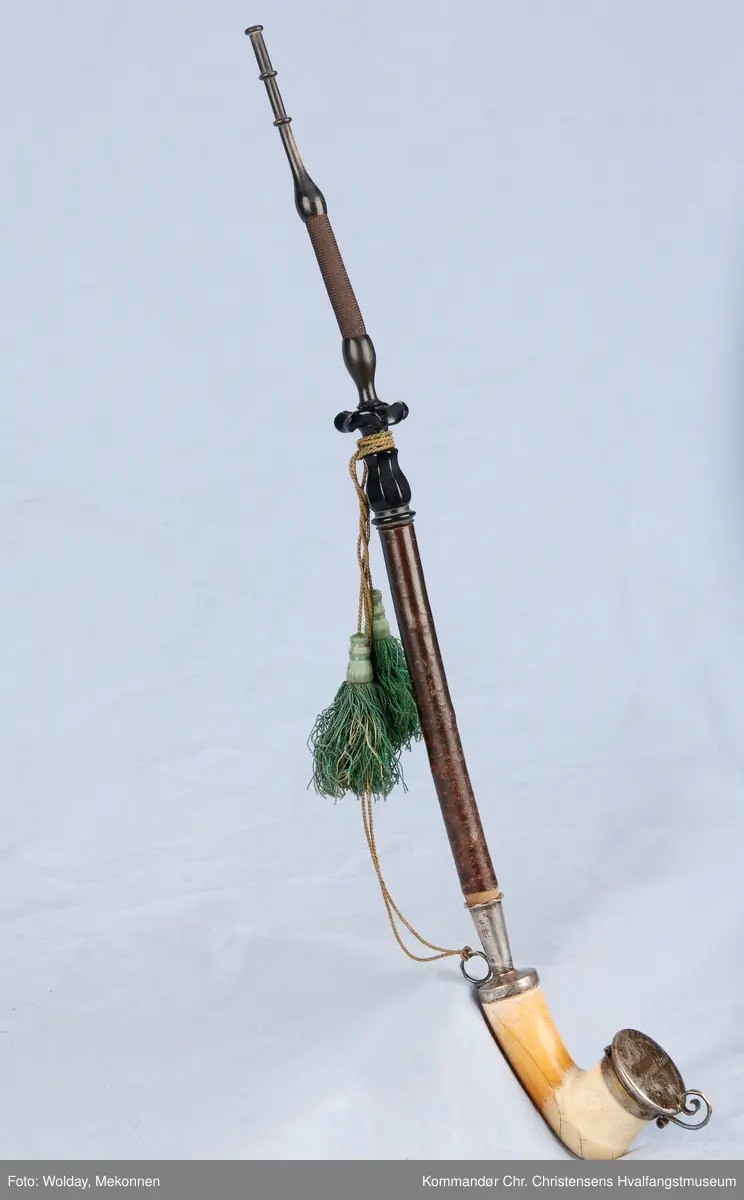 Langpipe med mellomstykke (holk) og lokk i sølv. Grønn snor med to dusker.