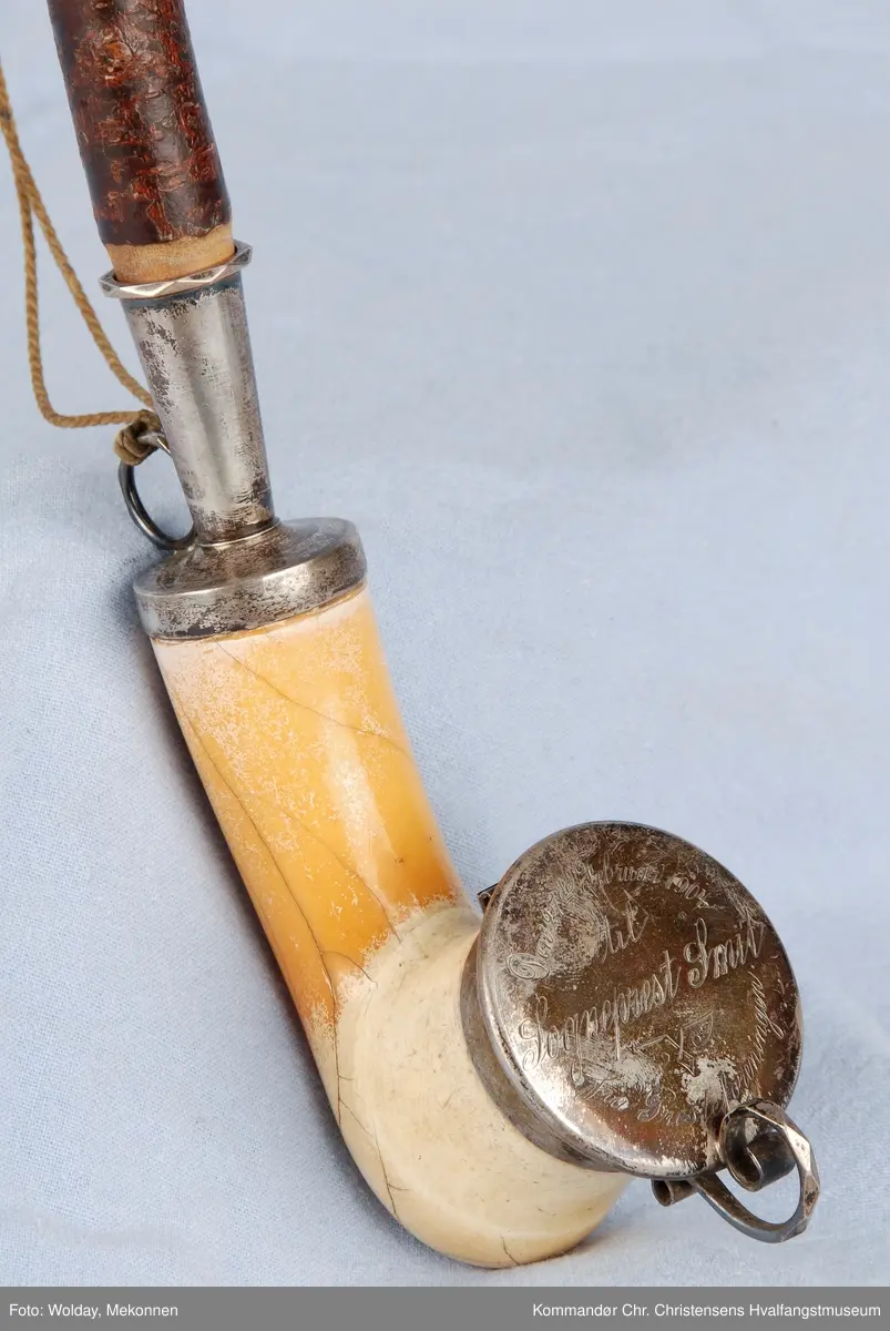 Langpipe med mellomstykke (holk) og lokk i sølv. Grønn snor med to dusker.