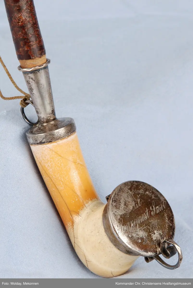 Langpipe med mellomstykke (holk) og lokk i sølv. Grønn snor med to dusker.