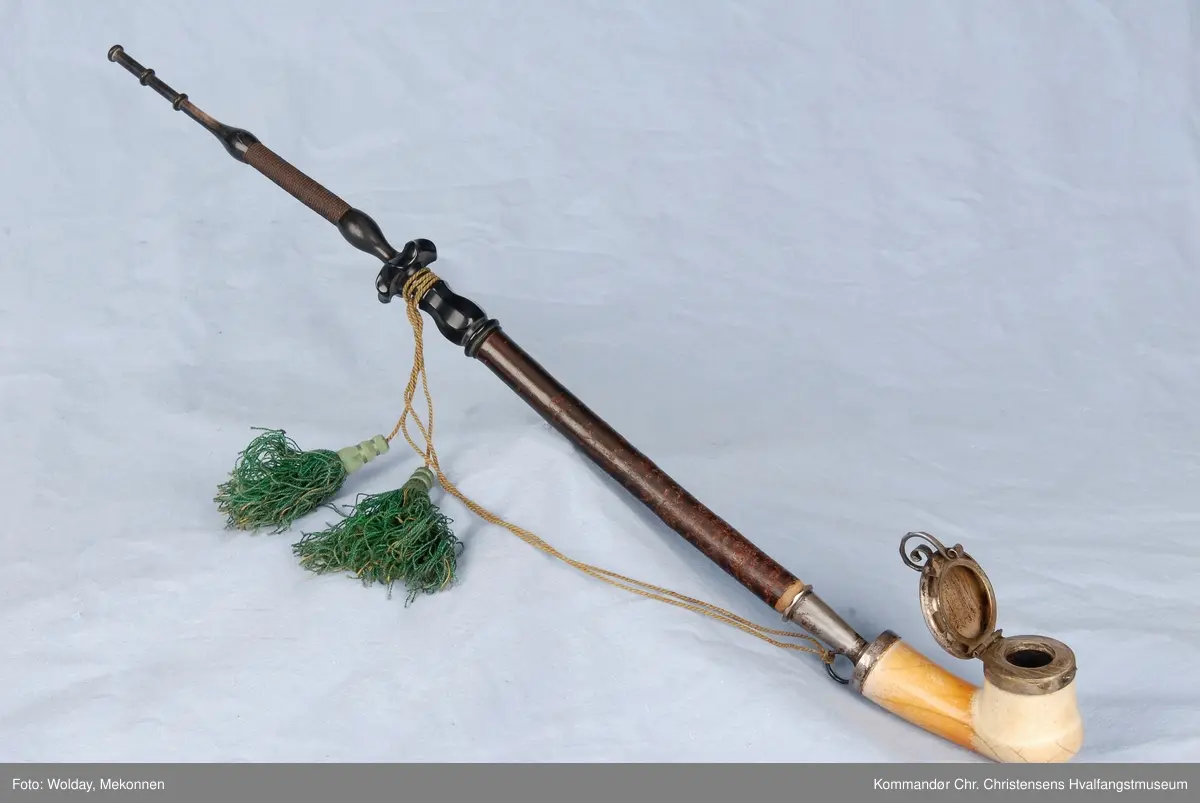 Langpipe med mellomstykke (holk) og lokk i sølv. Grønn snor med to dusker.