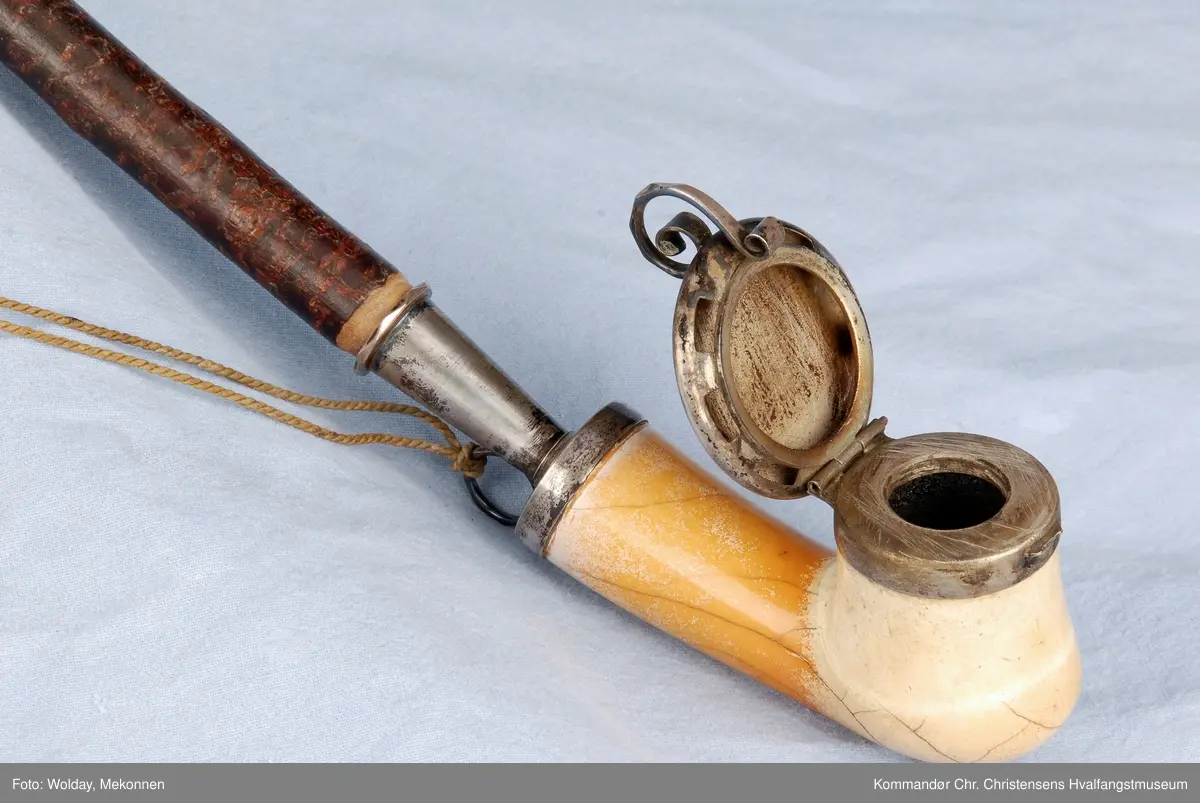 Langpipe med mellomstykke (holk) og lokk i sølv. Grønn snor med to dusker.