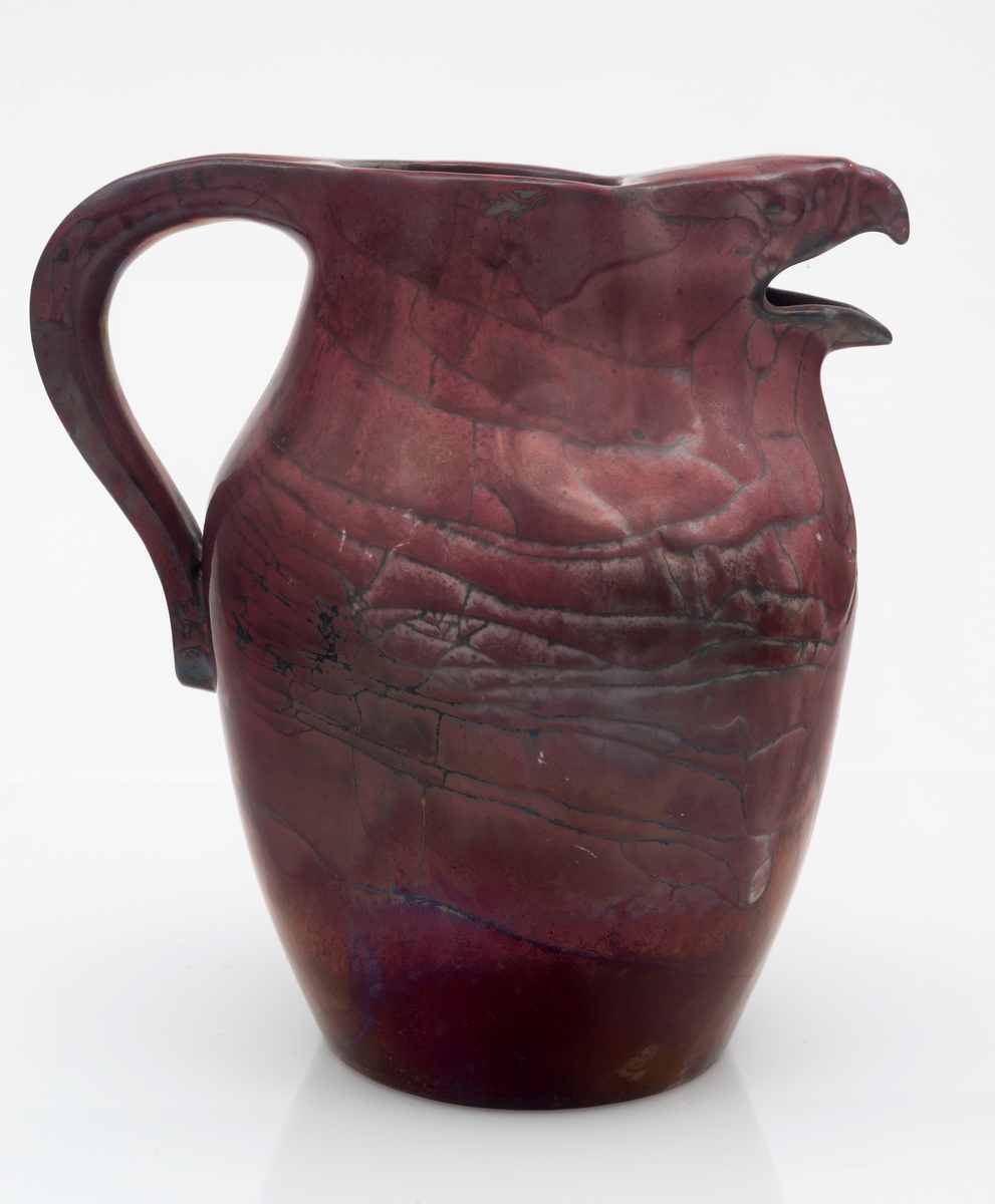 Kanne i glasert leirgods med hank. Tilnærmet ovalformet korpus med sirkulær åpning, hvor kannens tut er utformet som et skulpturelt haukehode. Overflaten er dekket med en profyrfarget glasur med store karakeleringer, hvor enkelte partier er irisert mens andre er hvite.