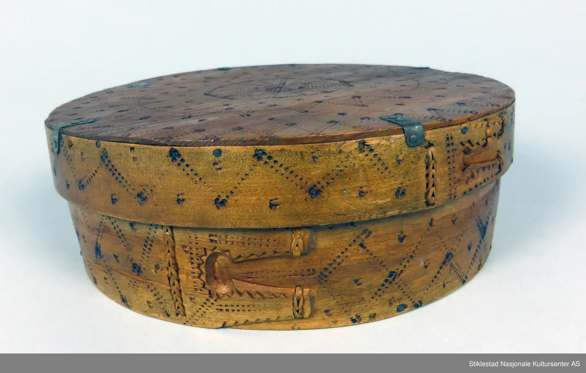 Sveipet øskje med søm av bjørketæger. Innbrent dekor på lokk og sider, rutemønster på side og lokk, 8-kant stjerne. Lokket er forsterket med vinkler av jernblikk. Trenaglene er erstatt med jernstifter
