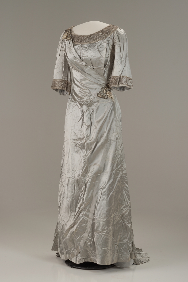 Skreddersydd, sølvfarget selskapskjole i kunstsilke med halvlange ermer og slep bak. Åpen, rund hals kantet med perlebroderi. Perlebroderi også nederst på ermene og til venstre i midjen. Flettet bån i kjolens stoff i ryggen. Øvre del av kjolen og ermene er fôret med bomull med isydde spiler av metall. Fôret lukkes med hekter. I fronten festes fôret i kjolen med trykknapper.