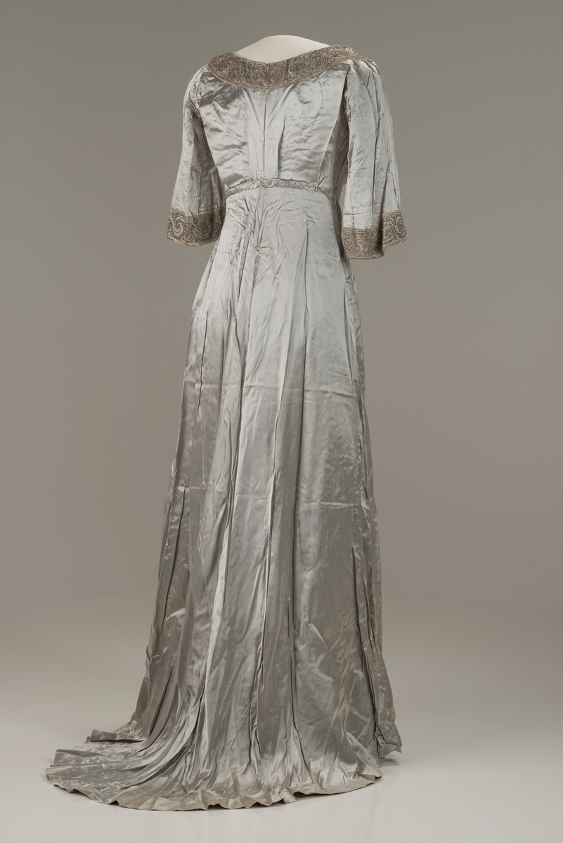 Skreddersydd, sølvfarget selskapskjole i kunstsilke med halvlange ermer og slep bak. Åpen, rund hals kantet med perlebroderi. Perlebroderi også nederst på ermene og til venstre i midjen. Flettet bån i kjolens stoff i ryggen. Øvre del av kjolen og ermene er fôret med bomull med isydde spiler av metall. Fôret lukkes med hekter. I fronten festes fôret i kjolen med trykknapper.