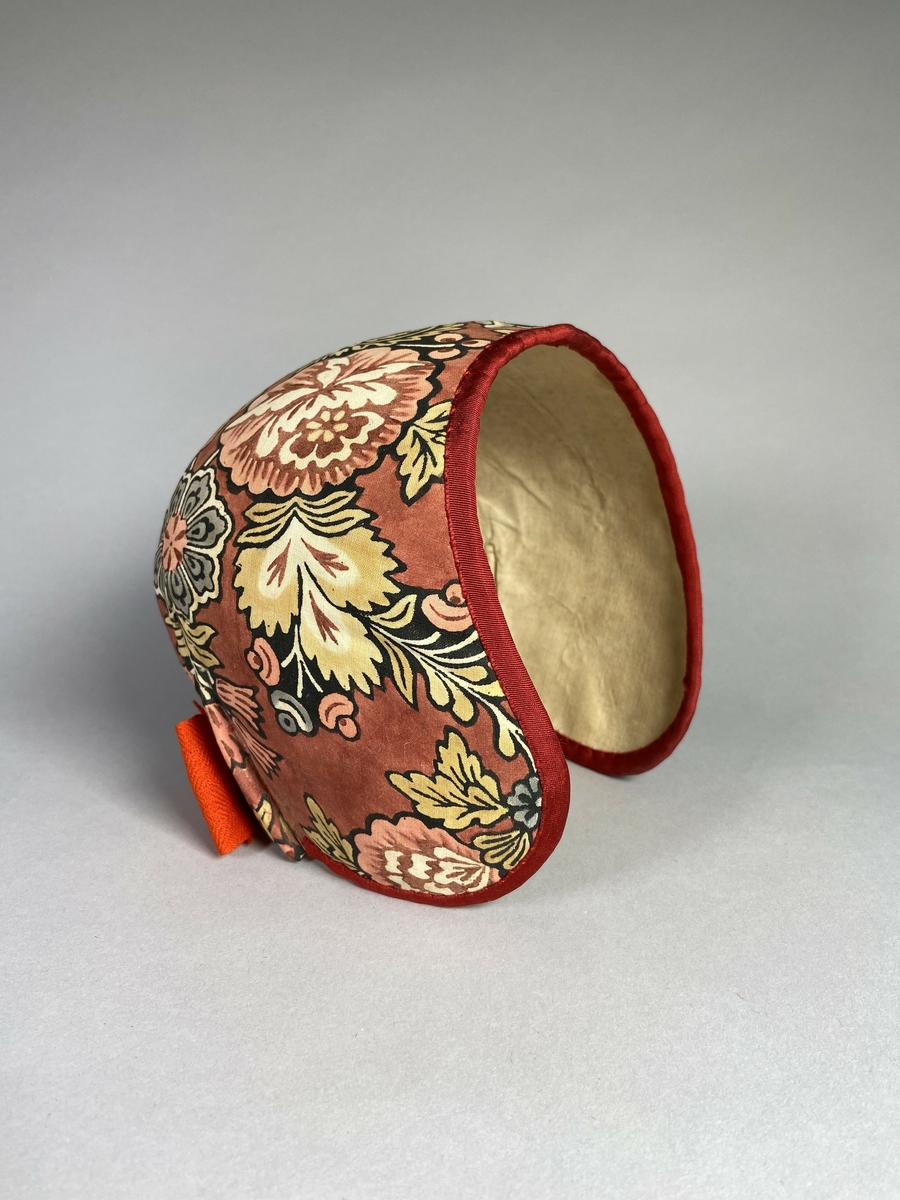 Bindmössa till Delsbodräkten. Klädd med tryckt och bemålad kattun med blomstermönster på röd botten. Stomme av papper och tyg. Kanter klädda med rött sidenband.
