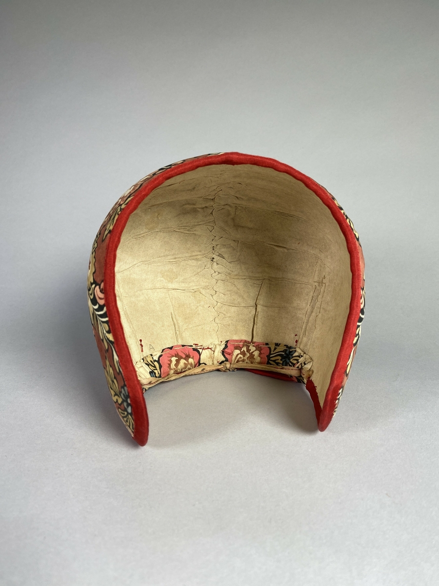 Bindmössa till Delsbodräkten. Klädd med tryckt och bemålad kattun med blomstermönster på röd botten. Stomme av papper och tyg. Kanter klädda med rött sidenband.
