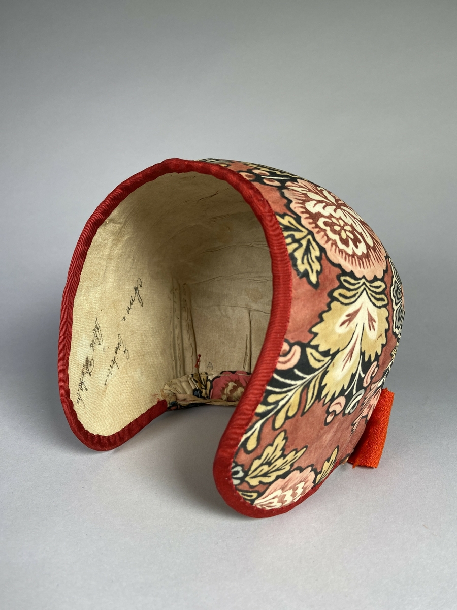 Bindmössa till Delsbodräkten. Klädd med tryckt och bemålad kattun med blomstermönster på röd botten. Stomme av papper och tyg. Kanter klädda med rött sidenband.
