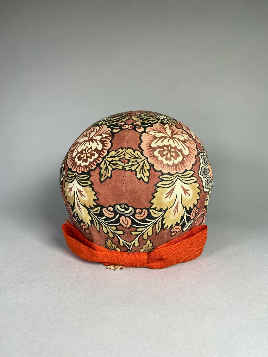 Bindmössa till Delsbodräkten. Klädd med tryckt och bemålad kattun med blomstermönster på röd botten. Stomme av papper och tyg. Kanter klädda med rött sidenband.
