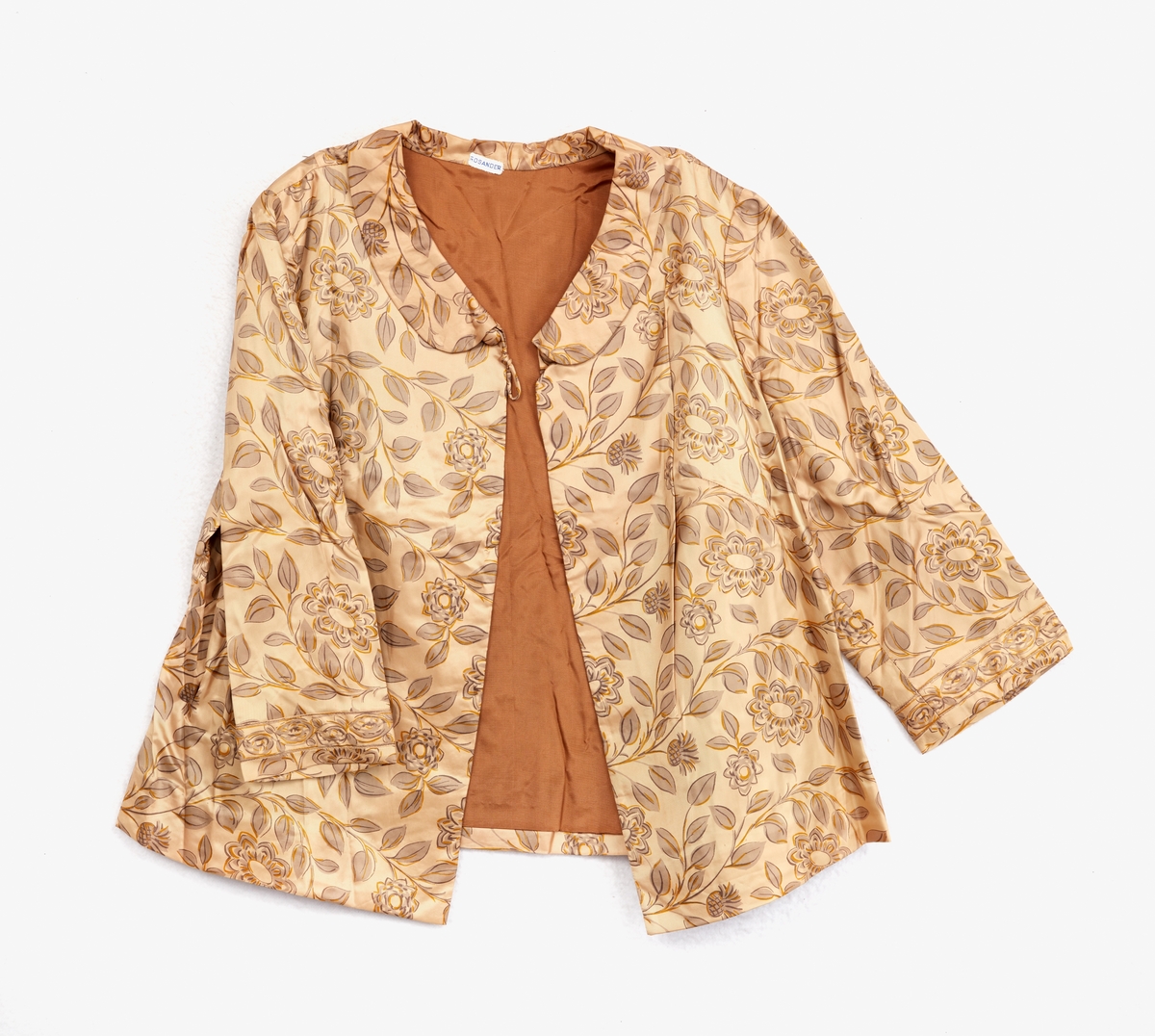 Klänning, ärmlös. Av ljusbrunt blankt tyg med tryckt mönster, blommor och blad. Kvadratisk halsringning. Dragkedja mitt bak, rosett vid halsringningen.
Underkjol med klänningstyget nedtill. 
Jacka av samma tyg som klänningen. Krage. 3/4 ärm. En knapp vid kragen. Baktill två löst hängande tygstycken, 'skört'. Fodrad med ljusbrunt tyg.
Märkt: 'Rosander'.