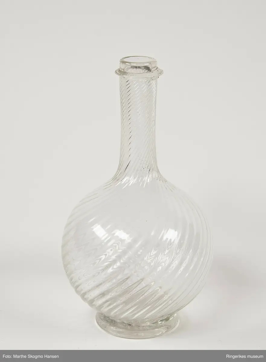 Glassflaske med spiralformede relieff linjer lang og smal hals, sirkelformet kropp og en liten stett.