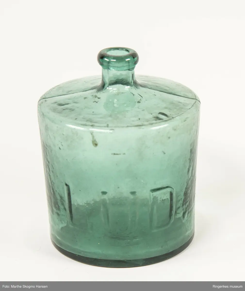 Grønn rund glassflaske med kort hals. Ordet `LUD` i utheva relieff.
