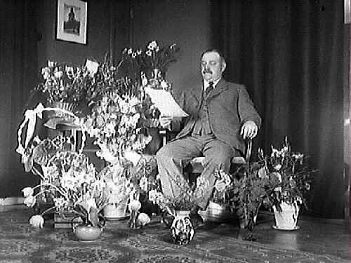 Maskinist Persson på Varbergs sjukhus fotograferad på sin 50 års-dag bland alla gratulationsblomster. Troligen är det ett telegram han håller i handen.