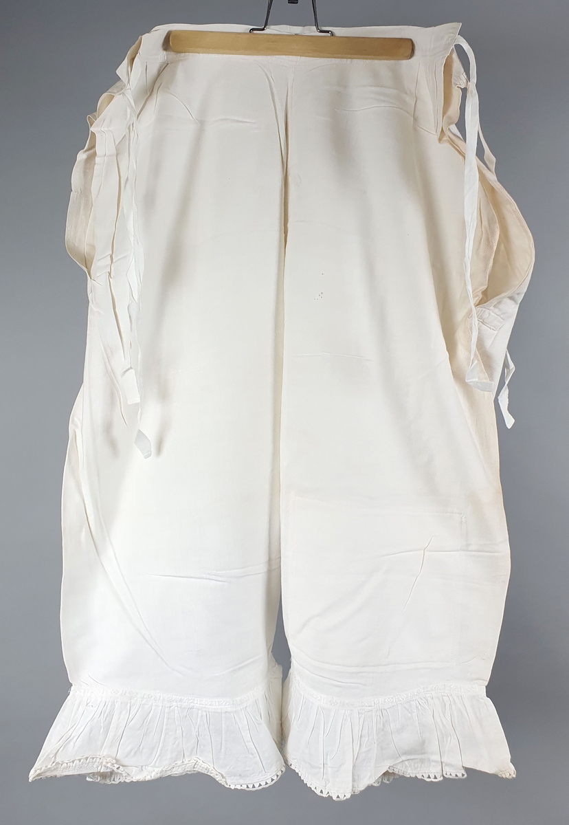 Ni like mamelukker med foldekant med bølget kant nederst.  Åpning i hver side med knytebånd.