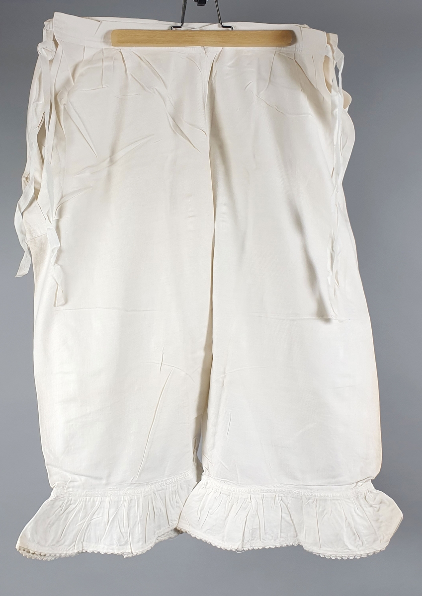 Ni like mamelukker med foldekant med bølget kant nederst.  Åpning i hver side med knytebånd.