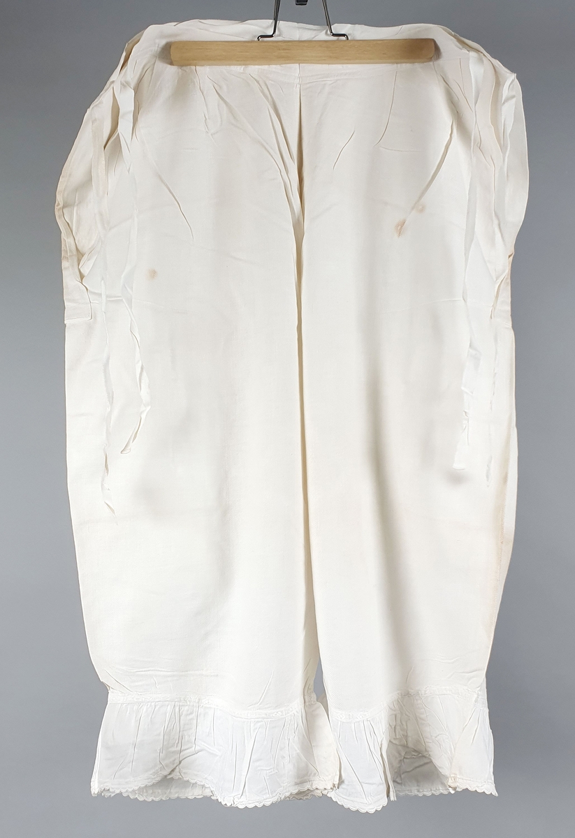 Ni like mamelukker med foldekant med bølget kant nederst.  Åpning i hver side med knytebånd.