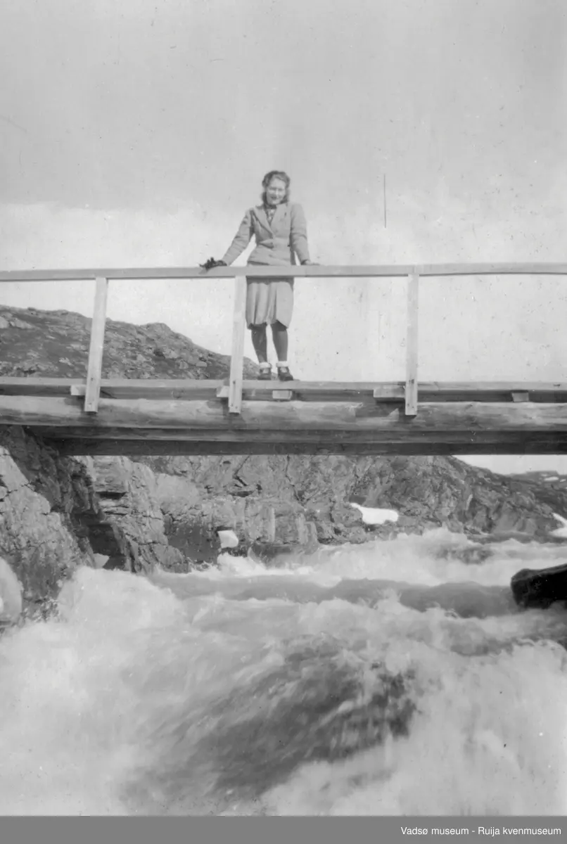 Ung kvinne oppstilt på trebro ovenfor foss. I bakgrunnen ses elva før fossen og steinete landskap.