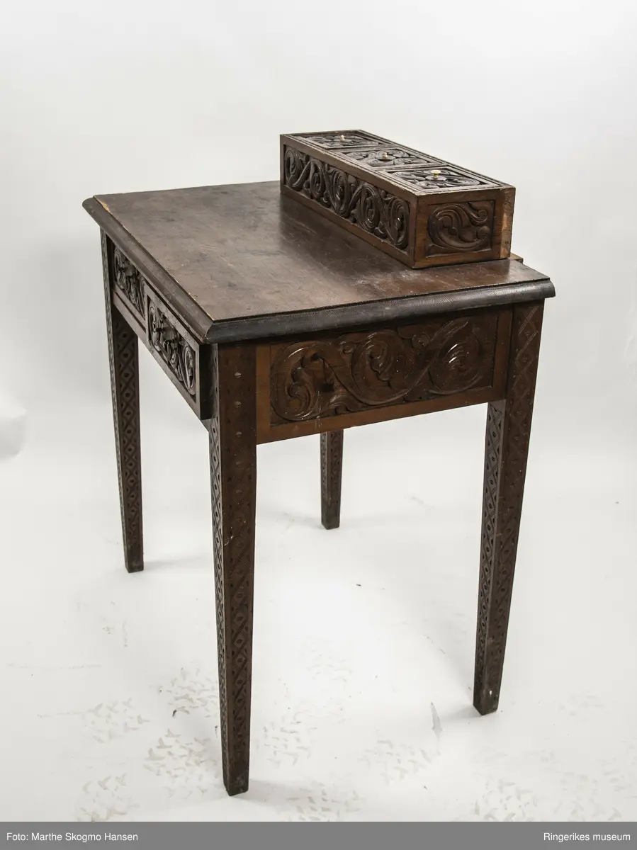 LIte bord med to skuffer og en boks på toppen. Fronten på skuffene, sidene på bordet, og boksen på toppen er skåret ut i tradisjonelt norsk akantusranke-mønster. Beina er skåre ut med geometriske mønstre på alle sider. Bordplata har en øvre del med rette kanter, men så buer den seg utover, så undersiden av bordet er større enn oversiden. Skuffene har håndtak av metall. Boksen er delt i tre rom med lokk. Lokkene er også skåret ut i samme mønster. De løftes ved hjelp av små håndtak, muligens av elfenben. To av disse er ødelagte. Boksen har helt tydelig vært festet i bordet, det er spor etter festene på hver side av boksen, og på baksiden av bordet er det skåret inn to hakk, et på hver side av boksen, disse er ca. 7 cm brede. I hver av disse hakkene står en bøyd, rusten spiker. På høyre side, sett forfra, har den biten av bordet som skulle vært på utsiden av hakket forsvunnet. Siden festet har blitt borte, er boksen nå løs. 