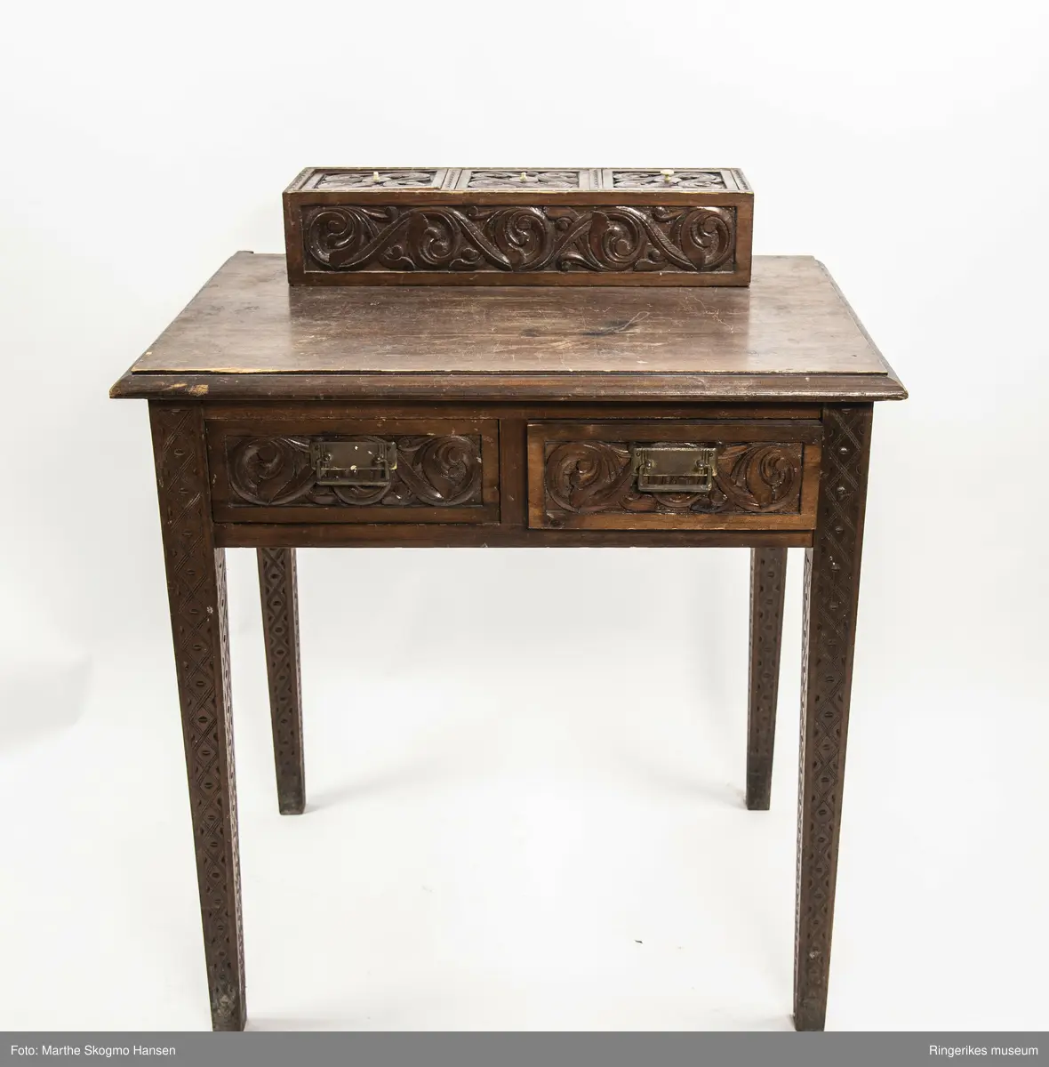 LIte bord med to skuffer og en boks på toppen. Fronten på skuffene, sidene på bordet, og boksen på toppen er skåret ut i tradisjonelt norsk akantusranke-mønster. Beina er skåre ut med geometriske mønstre på alle sider. Bordplata har en øvre del med rette kanter, men så buer den seg utover, så undersiden av bordet er større enn oversiden. Skuffene har håndtak av metall. Boksen er delt i tre rom med lokk. Lokkene er også skåret ut i samme mønster. De løftes ved hjelp av små håndtak, muligens av elfenben. To av disse er ødelagte. Boksen har helt tydelig vært festet i bordet, det er spor etter festene på hver side av boksen, og på baksiden av bordet er det skåret inn to hakk, et på hver side av boksen, disse er ca. 7 cm brede. I hver av disse hakkene står en bøyd, rusten spiker. På høyre side, sett forfra, har den biten av bordet som skulle vært på utsiden av hakket forsvunnet. Siden festet har blitt borte, er boksen nå løs. 