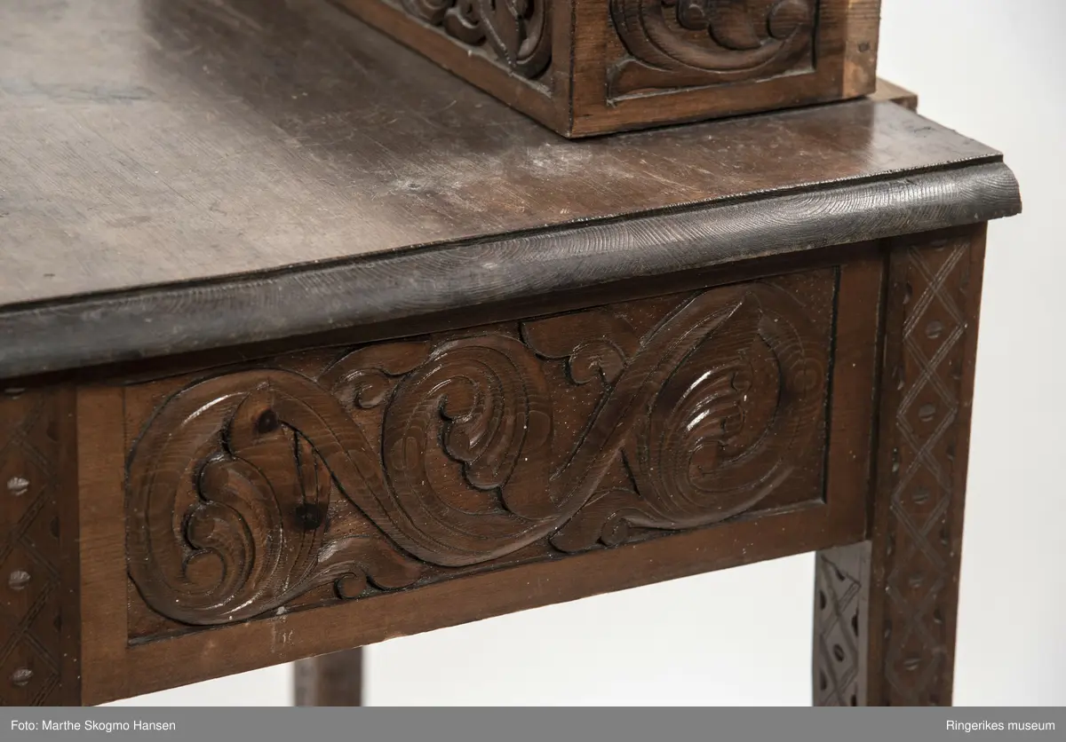 LIte bord med to skuffer og en boks på toppen. Fronten på skuffene, sidene på bordet, og boksen på toppen er skåret ut i tradisjonelt norsk akantusranke-mønster. Beina er skåre ut med geometriske mønstre på alle sider. Bordplata har en øvre del med rette kanter, men så buer den seg utover, så undersiden av bordet er større enn oversiden. Skuffene har håndtak av metall. Boksen er delt i tre rom med lokk. Lokkene er også skåret ut i samme mønster. De løftes ved hjelp av små håndtak, muligens av elfenben. To av disse er ødelagte. Boksen har helt tydelig vært festet i bordet, det er spor etter festene på hver side av boksen, og på baksiden av bordet er det skåret inn to hakk, et på hver side av boksen, disse er ca. 7 cm brede. I hver av disse hakkene står en bøyd, rusten spiker. På høyre side, sett forfra, har den biten av bordet som skulle vært på utsiden av hakket forsvunnet. Siden festet har blitt borte, er boksen nå løs. 