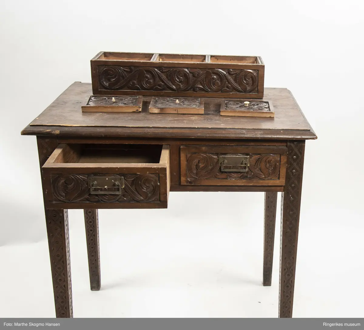 LIte bord med to skuffer og en boks på toppen. Fronten på skuffene, sidene på bordet, og boksen på toppen er skåret ut i tradisjonelt norsk akantusranke-mønster. Beina er skåre ut med geometriske mønstre på alle sider. Bordplata har en øvre del med rette kanter, men så buer den seg utover, så undersiden av bordet er større enn oversiden. Skuffene har håndtak av metall. Boksen er delt i tre rom med lokk. Lokkene er også skåret ut i samme mønster. De løftes ved hjelp av små håndtak, muligens av elfenben. To av disse er ødelagte. Boksen har helt tydelig vært festet i bordet, det er spor etter festene på hver side av boksen, og på baksiden av bordet er det skåret inn to hakk, et på hver side av boksen, disse er ca. 7 cm brede. I hver av disse hakkene står en bøyd, rusten spiker. På høyre side, sett forfra, har den biten av bordet som skulle vært på utsiden av hakket forsvunnet. Siden festet har blitt borte, er boksen nå løs. 