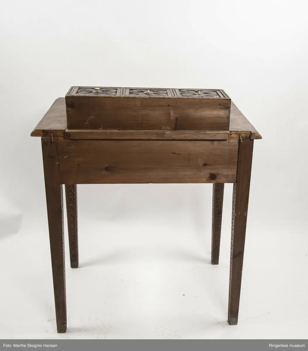 LIte bord med to skuffer og en boks på toppen. Fronten på skuffene, sidene på bordet, og boksen på toppen er skåret ut i tradisjonelt norsk akantusranke-mønster. Beina er skåre ut med geometriske mønstre på alle sider. Bordplata har en øvre del med rette kanter, men så buer den seg utover, så undersiden av bordet er større enn oversiden. Skuffene har håndtak av metall. Boksen er delt i tre rom med lokk. Lokkene er også skåret ut i samme mønster. De løftes ved hjelp av små håndtak, muligens av elfenben. To av disse er ødelagte. Boksen har helt tydelig vært festet i bordet, det er spor etter festene på hver side av boksen, og på baksiden av bordet er det skåret inn to hakk, et på hver side av boksen, disse er ca. 7 cm brede. I hver av disse hakkene står en bøyd, rusten spiker. På høyre side, sett forfra, har den biten av bordet som skulle vært på utsiden av hakket forsvunnet. Siden festet har blitt borte, er boksen nå løs. 