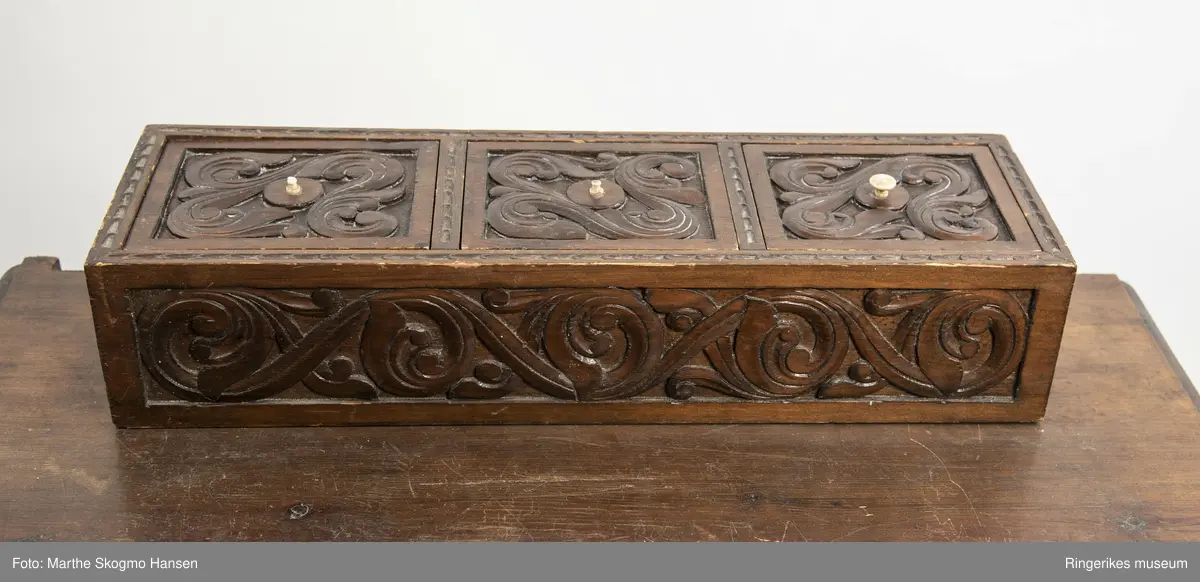 LIte bord med to skuffer og en boks på toppen. Fronten på skuffene, sidene på bordet, og boksen på toppen er skåret ut i tradisjonelt norsk akantusranke-mønster. Beina er skåre ut med geometriske mønstre på alle sider. Bordplata har en øvre del med rette kanter, men så buer den seg utover, så undersiden av bordet er større enn oversiden. Skuffene har håndtak av metall. Boksen er delt i tre rom med lokk. Lokkene er også skåret ut i samme mønster. De løftes ved hjelp av små håndtak, muligens av elfenben. To av disse er ødelagte. Boksen har helt tydelig vært festet i bordet, det er spor etter festene på hver side av boksen, og på baksiden av bordet er det skåret inn to hakk, et på hver side av boksen, disse er ca. 7 cm brede. I hver av disse hakkene står en bøyd, rusten spiker. På høyre side, sett forfra, har den biten av bordet som skulle vært på utsiden av hakket forsvunnet. Siden festet har blitt borte, er boksen nå løs. 