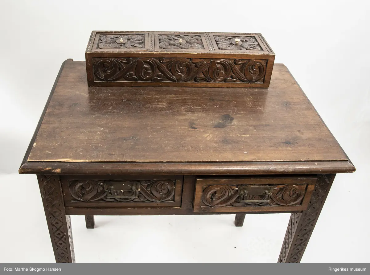 LIte bord med to skuffer og en boks på toppen. Fronten på skuffene, sidene på bordet, og boksen på toppen er skåret ut i tradisjonelt norsk akantusranke-mønster. Beina er skåre ut med geometriske mønstre på alle sider. Bordplata har en øvre del med rette kanter, men så buer den seg utover, så undersiden av bordet er større enn oversiden. Skuffene har håndtak av metall. Boksen er delt i tre rom med lokk. Lokkene er også skåret ut i samme mønster. De løftes ved hjelp av små håndtak, muligens av elfenben. To av disse er ødelagte. Boksen har helt tydelig vært festet i bordet, det er spor etter festene på hver side av boksen, og på baksiden av bordet er det skåret inn to hakk, et på hver side av boksen, disse er ca. 7 cm brede. I hver av disse hakkene står en bøyd, rusten spiker. På høyre side, sett forfra, har den biten av bordet som skulle vært på utsiden av hakket forsvunnet. Siden festet har blitt borte, er boksen nå løs. 