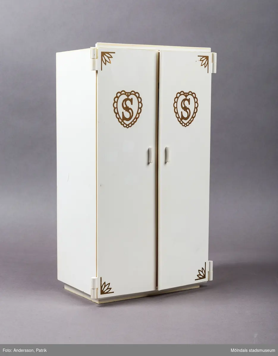 Vit leksaksgarderob med gulddekor. Till skåpet hör fem galgar och två papiljotter. 

Inuti garderoben finns en liten byrå med tre lådor (en låda saknas dock).  På insidan av den vänstra garderobsdörren finns en spegel och en skohylla. På den högra dörren finns en liten hylla och en stång för skärp och scarfs. 

Garderoben är ett tillbehör till dockan Sindy.

Sindy var en docka som tillverkades av ett stort antal företag på licens från Pedigree Dolls & Toys: Pedigree under åren 1963-1986, Hasbro 1987-1996, Funskool 1994-2006 samt Vivid Imaginations 1998-2003.