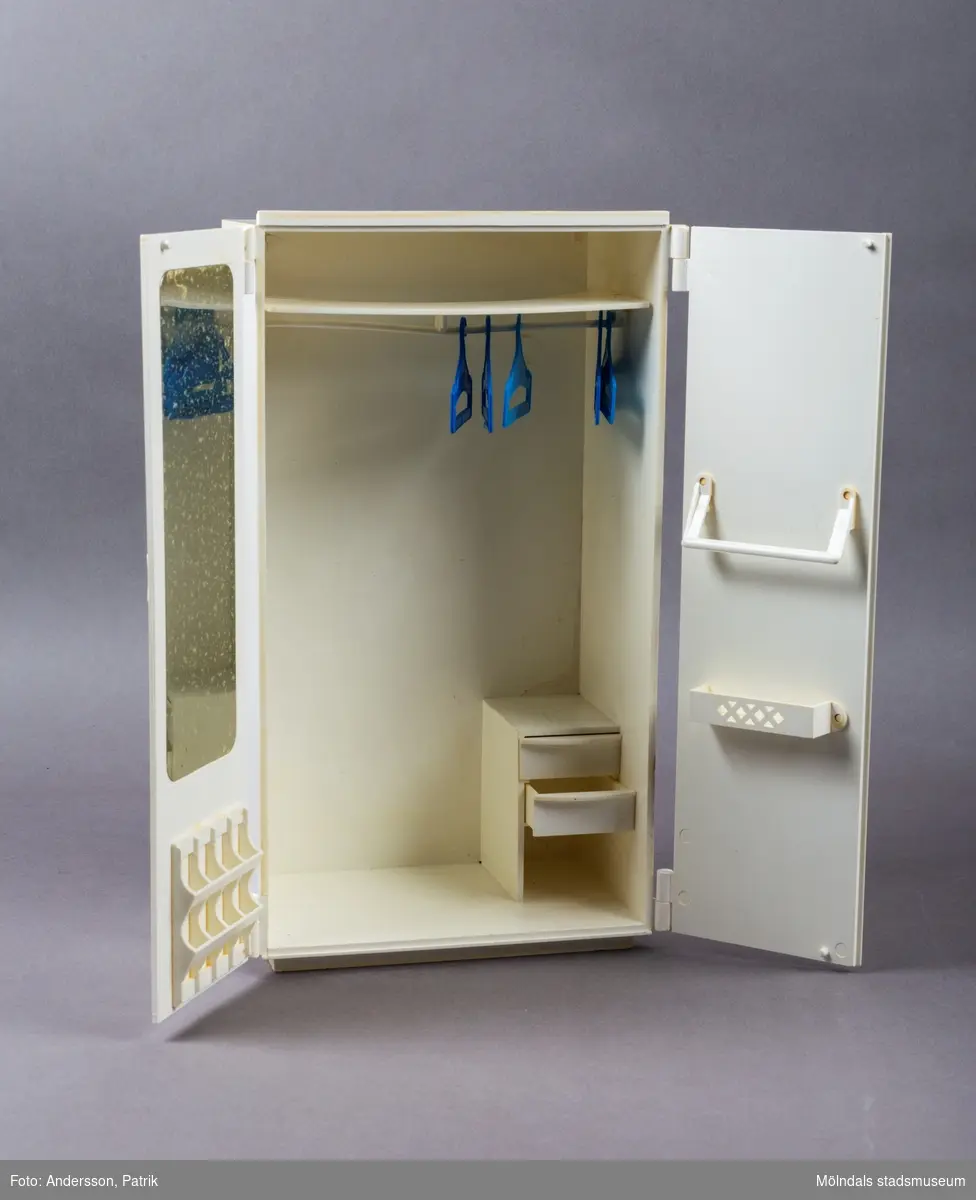 Vit leksaksgarderob med gulddekor. Till skåpet hör fem galgar och två papiljotter. 

Inuti garderoben finns en liten byrå med tre lådor (en låda saknas dock).  På insidan av den vänstra garderobsdörren finns en spegel och en skohylla. På den högra dörren finns en liten hylla och en stång för skärp och scarfs. 

Garderoben är ett tillbehör till dockan Sindy.

Sindy var en docka som tillverkades av ett stort antal företag på licens från Pedigree Dolls & Toys: Pedigree under åren 1963-1986, Hasbro 1987-1996, Funskool 1994-2006 samt Vivid Imaginations 1998-2003.