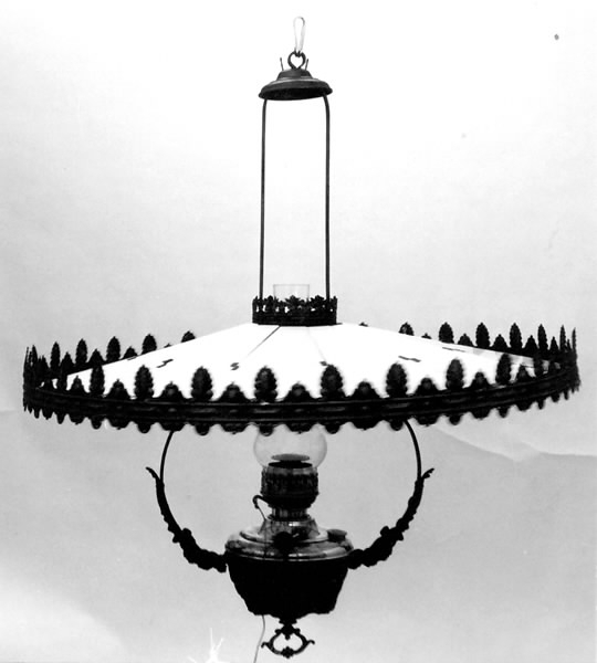 Rund lampskärm av plåt och glas. En yttre och inre cirkel med palmette- och rankdekor. Mjölkvitt glas i form av tårtbitar från yttre till inre cirkeln. Till fotogenlampa VMF 030896. Höjd: 12,5 cm. Diameter: 76 cm.