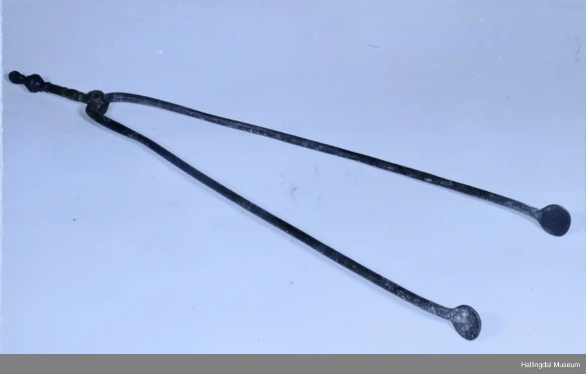 Bena har sirkulært tverrsnitt. Buer svakt innover mot hverandre og avsluttes i flatbanket, sirkulær plate. I den andre ende bøyd innover og festet sammen, slik at de kan dreies fra hverandre. Det ene ben fortsetter ut i håndtak, sirkulært tverrsnitt. Profiler og vulst. Ender i en liten løkke.
25/6-67 O. Foldøy.