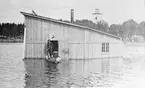 Löjtnant Tor von Johnstone står i en båt vid en hangar vid gamla flygstationen vid Bodträsket. Överluleå kyrka i bakgrunden. Troligen vid översvämning, 1917.