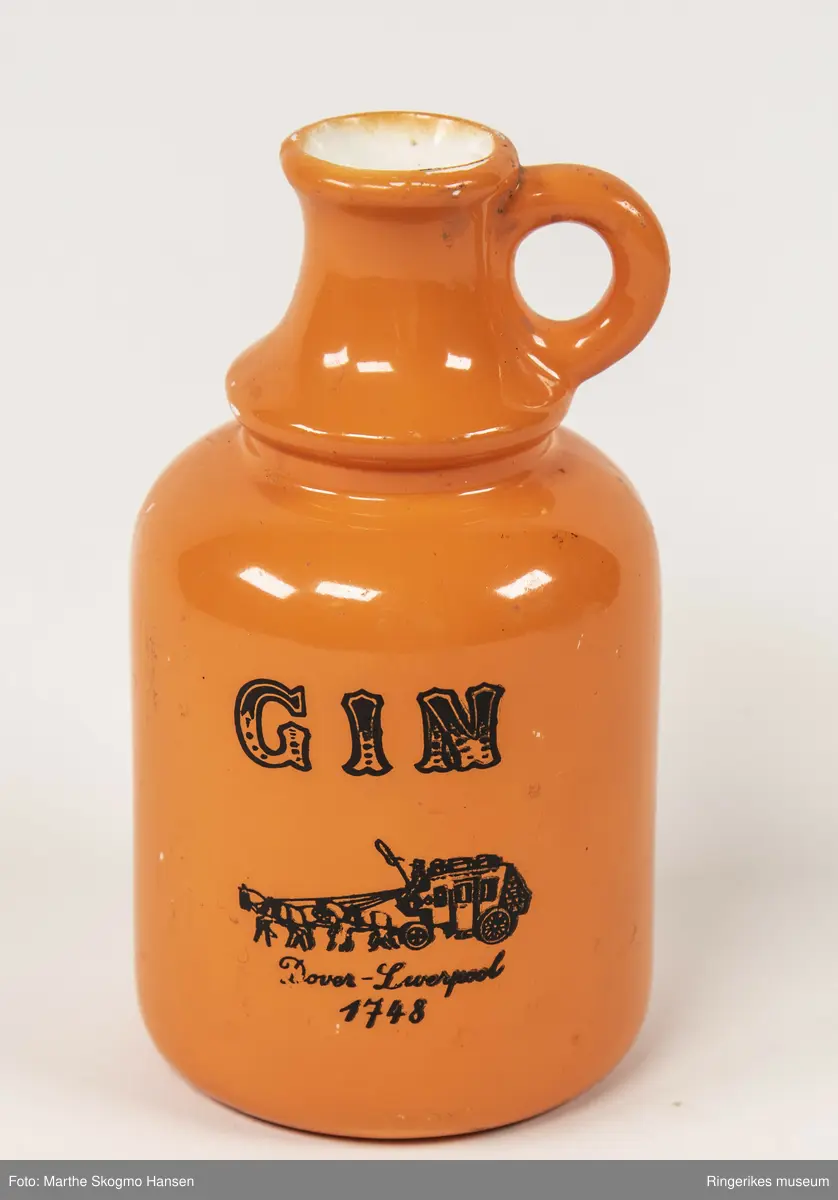 Orange keramikkflaske med en liten rund hank øverst på halsen. Flaska har hvit innside. Foran på flaska står det GIN Dover-Liverpool 1748 og en tegning av to hester som trekker en vogn.