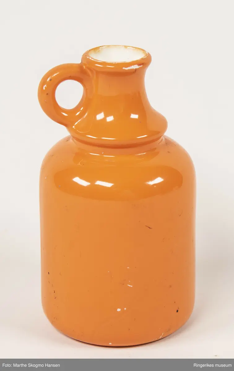 Orange keramikkflaske med en liten rund hank øverst på halsen. Flaska har hvit innside. Foran på flaska står det GIN Dover-Liverpool 1748 og en tegning av to hester som trekker en vogn.