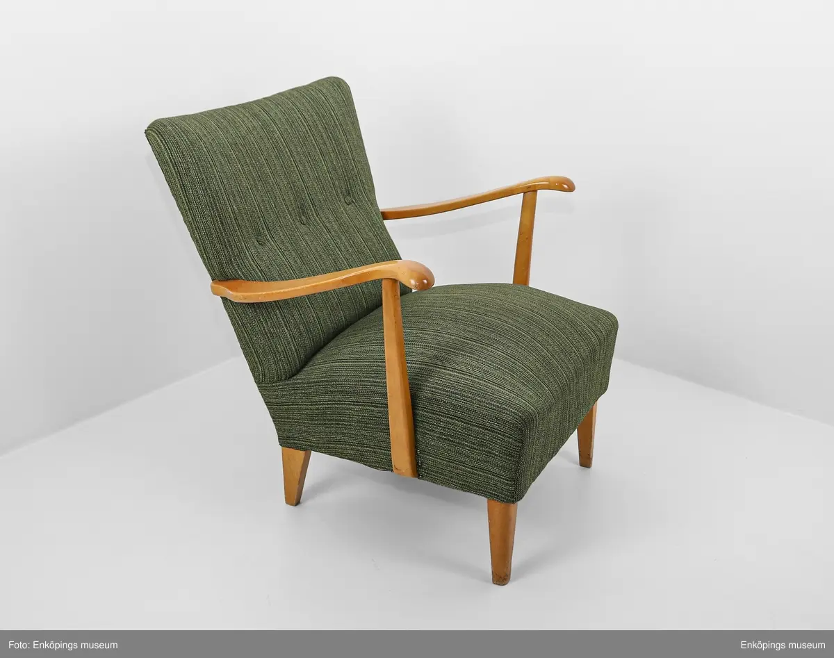 Grön fåtölj från 1950-talet. Ljust träslag i armstöd och ben. Sits och ryggstöd klädda med vävt tyg. På stolsryggens framsida finns tre textilklädda knappar.