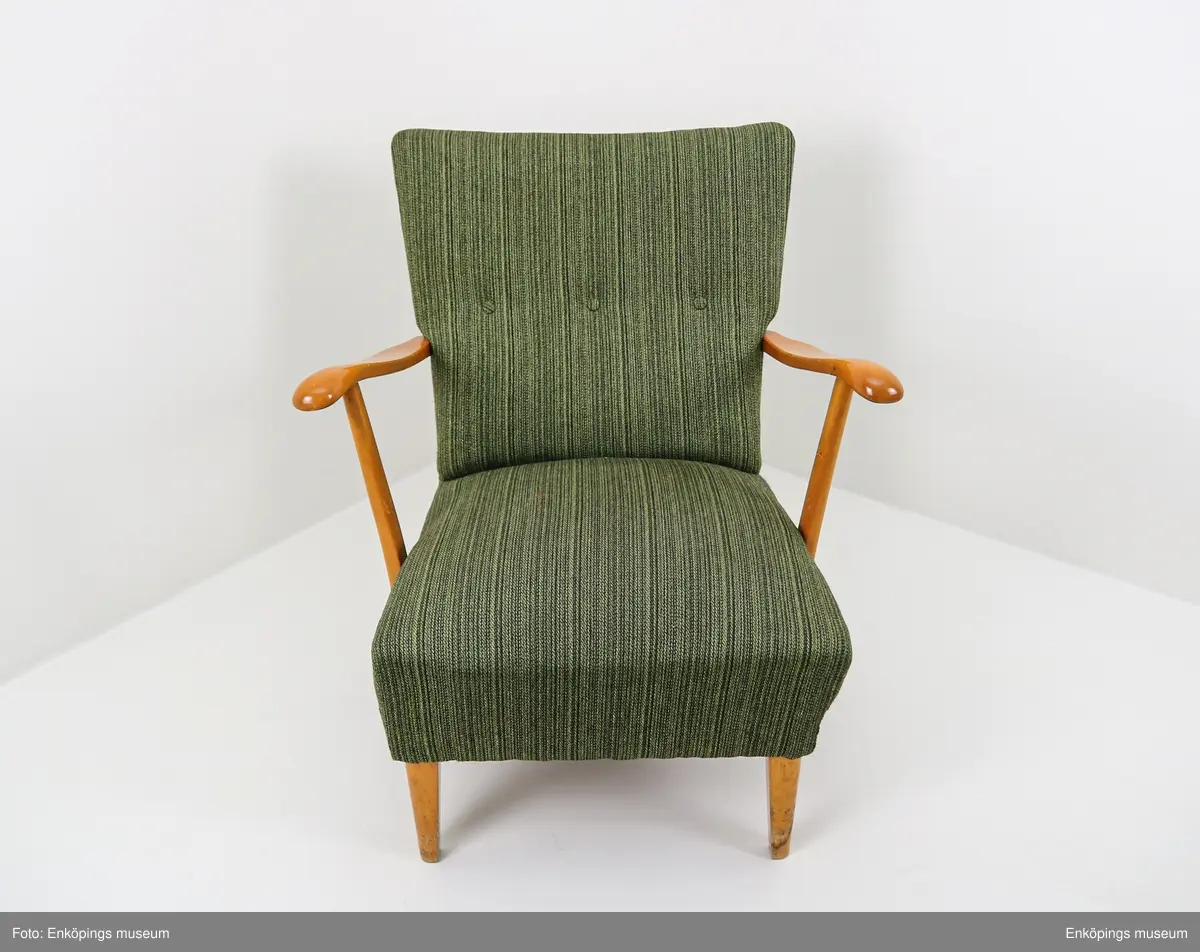 Grön fåtölj från 1950-talet. Ljust träslag i armstöd och ben. Sits och ryggstöd klädda med vävt tyg. På stolsryggens framsida finns tre textilklädda knappar.