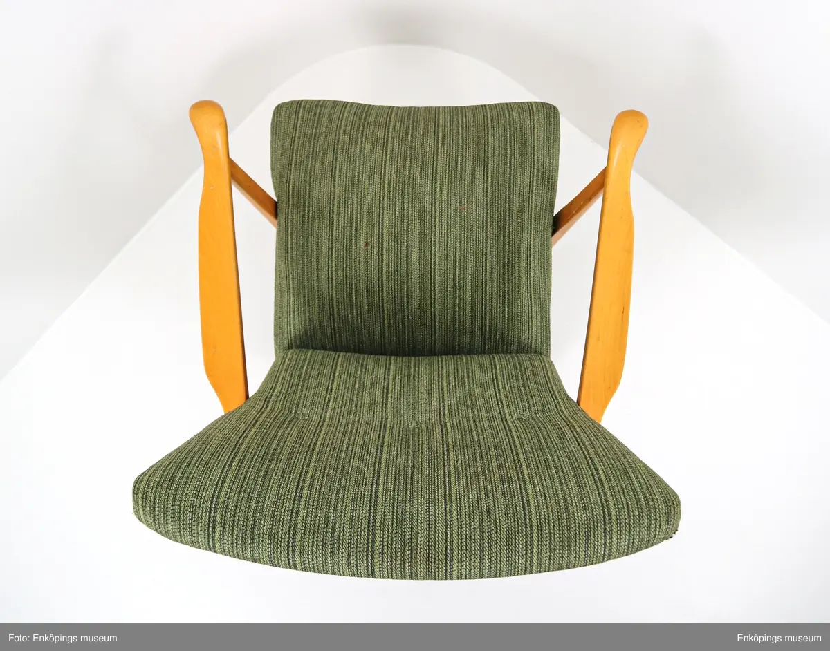 Grön fåtölj från 1950-talet. Ljust träslag i armstöd och ben. Sits och ryggstöd klädda med vävt tyg. På stolsryggens framsida finns tre textilklädda knappar.