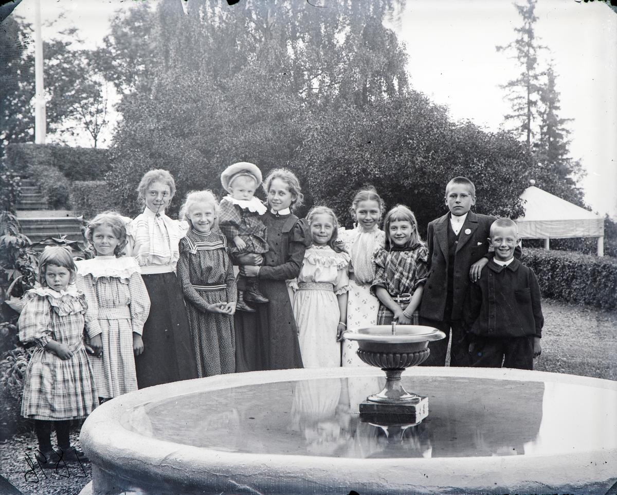 Barn rundt fontene i hagen på Hovinsholm 1898: Fra v.: Søstrene Thora Hoel og Martha Louise Hoel, Bergljot Rogneby, Bergljot Bassøe, Helga Sandberg med Reidar Ramm Boe, Ragna Møller, Lilly Meyer, Ambjørg Rogneby, Gustav Hoel, Einar Møller.