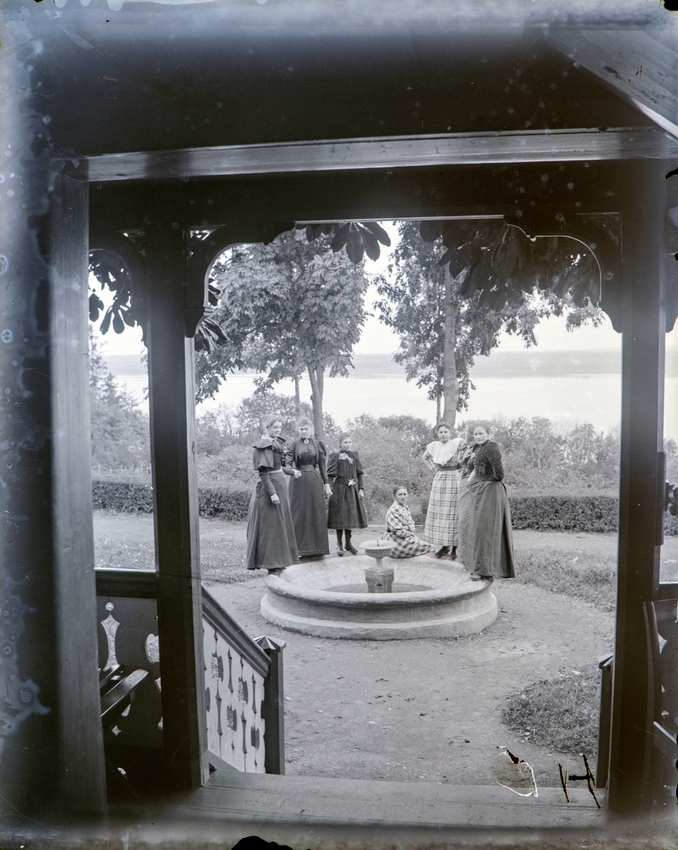 Bilde tatt innen fra, og ut mot på Hovinsholm: Agnes Olsen, ukjent, Sigrid Pauline, Albertine, Ragnhild Marie og kusine Marie Ramm Hoel er samlet rundt vannfontenen i hagen.