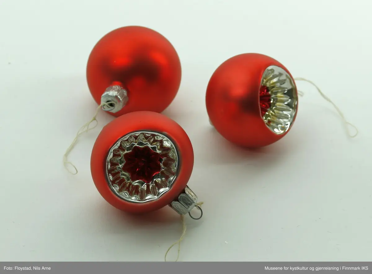 Tre røde julekuler med mønsterformet uthuling på framsiden av kulene. De er alle små i størrelse, men det er en veldig liten forskjell på størrelsen på alle tre. Alle kulene er laget av plastmateriale. Kulene har en halvmatt lysrød farge. Uthulingen på kulene har et stjerneformet mønster som går inn til midten av kulene. Mønsteret har en blank sølvfarge i ytterkanten og i midten har det en blank mørkerødfarge. Helt i midten har mønsteret på uthulingene form som en mørkerød stjerne. Kulene har oppheng i metall med gravert mønsterdekor, som består av små spor og mønster formet som et 8-tall. Opphengene har en liten tråd knytt på.