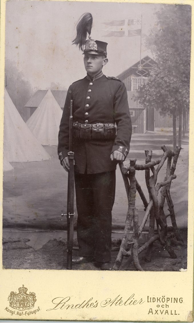 Kabinettsfotografi av en okänd ung man i militär uniform. Han står med ett gevär intill sig mot en fond av militär bivack.