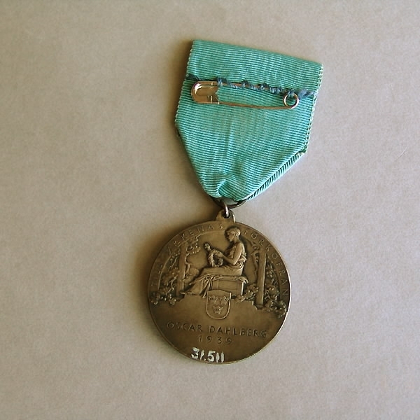 Rund medalj, försilvrad. Diameter 4 cm. Medaljen är fastsatt i ett blått sidenband. Framsida: Gustav V i profil samt text: "GUSTAV V SVERIGES HANTVERKSORGANISATIONS BESKYDDARE". Baksida: Kvinna knytande en krans, sittande på en tron. En sköld med tre kronor samt text: "TILL YRKENAS FÖRKOFRAN OSCAR DAHLBERG 1939". En säkerhetsnål fastsydd på sidenbandets baksida.