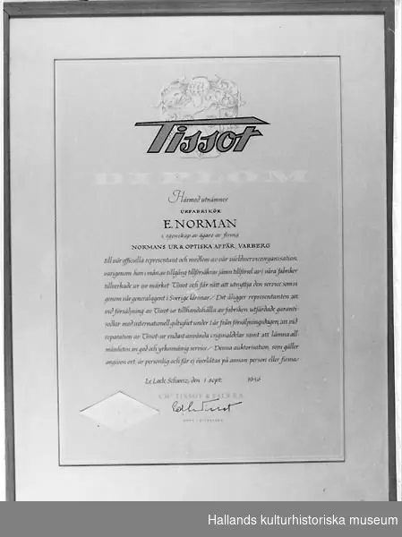 Diplom med träram (bredd= 1 cm) och glas. Ståltråd för upphängning baktill.Överst Tissots firmamärke. Text: "Tissot DIPLOM" "Härmed utnämnes URFABRIKÖR E. NORMAN i egenskap av ägare av firma NORMANS UR & OPTISKA AFFÄR, VARBERG till representant och medlem av vår världsserviceorganisation, varigenom han i mån av tillgång tillförsäkras jämn tillförsel av i våra fabriker tillverkade ur av märket Tissot och får rätt att utnyttja den service som vi genom vår generalagent i Sverige lämnar/ Det åligger representanten att vid försäljning av Tissot-ur tillhandahålla av fabriken utfärdade garantisedlar med internationell giltighet under 1 år från försäljningsdagen vid reparation av Tissot-ur endast använda originaldelar samt att lämna en god och yrkesmässig service/ Denna auktorisation, som gäller angiven ort, är personlig och får ej överlåtas på annan person eller firma/ Le Loche, Schweiz den 1 sept. 1956" CHs TISSOT & FILS S.A." Undertecknat av ? ? "ORDF I STYRELSEN."