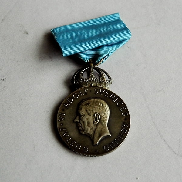 Bronsmedalj med förvaringsask av papp. a) Medalj av brons. Märkt på framsidan: "Gustav VI Adolf Sveriges Konung". Samt profil av huvudet. Baksidan märkt: "Sveriges hantverks- och småindustri organisation. J. Carlsson 1954". Blått sidenband med säkerhetsnål. Höjd 10 cm. Bredd 4 cm. Tjocklek 0,5 cm. b) Ask som medaljen ligger i. Blå, av papp med blå sammet på insidan. Diameter b):100 mm. Bredd b): 60 mm. Höjd b): 20 mm.