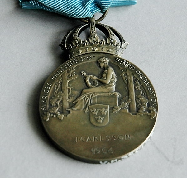 Bronsmedalj med förvaringsask av papp. a) Medalj av brons. Märkt på framsidan: "Gustav VI Adolf Sveriges Konung". Samt profil av huvudet. Baksidan märkt: "Sveriges hantverks- och småindustri organisation. J. Carlsson 1954". Blått sidenband med säkerhetsnål. Höjd 10 cm. Bredd 4 cm. Tjocklek 0,5 cm. b) Ask som medaljen ligger i. Blå, av papp med blå sammet på insidan. Diameter b):100 mm. Bredd b): 60 mm. Höjd b): 20 mm.